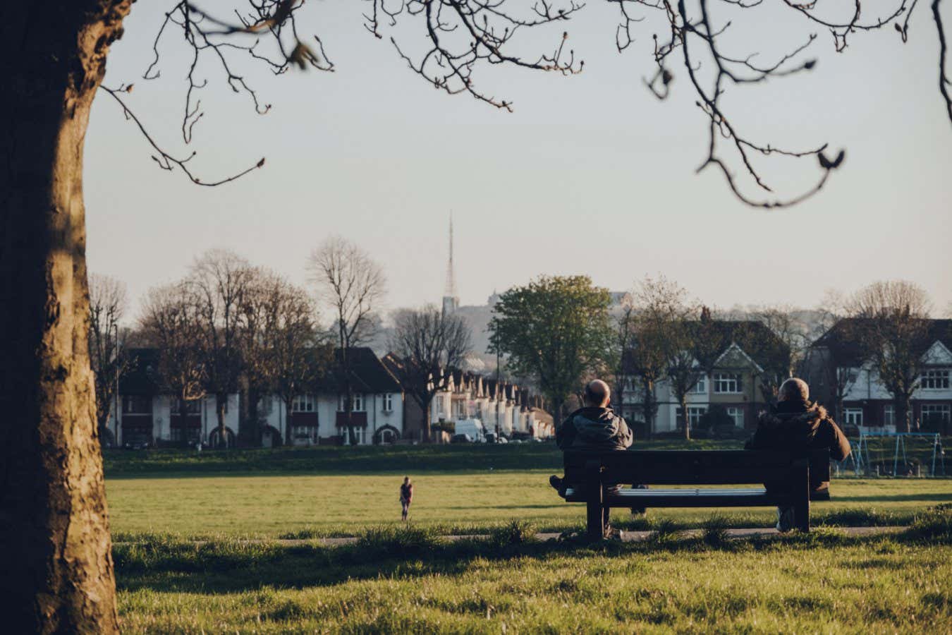 2BCPF0B London, UK - March 22,2020: Rear view of two men sitting on the opposite sides of the bench watching people play ball in Broomfield Park, public park